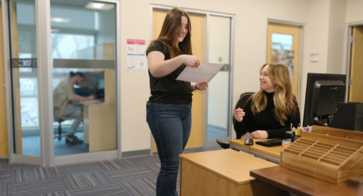 Student holding document standing in front of person sitting at desk