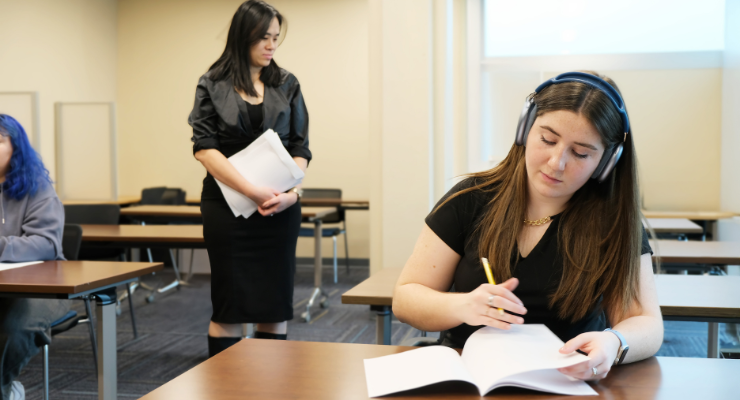 Student wearing headphones writing test at desk while invigilator walks by