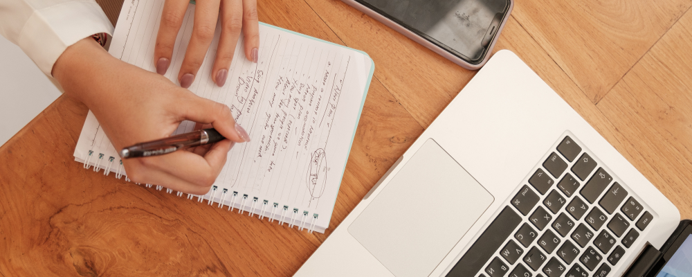 A laptop and hand taking notes in a notebook