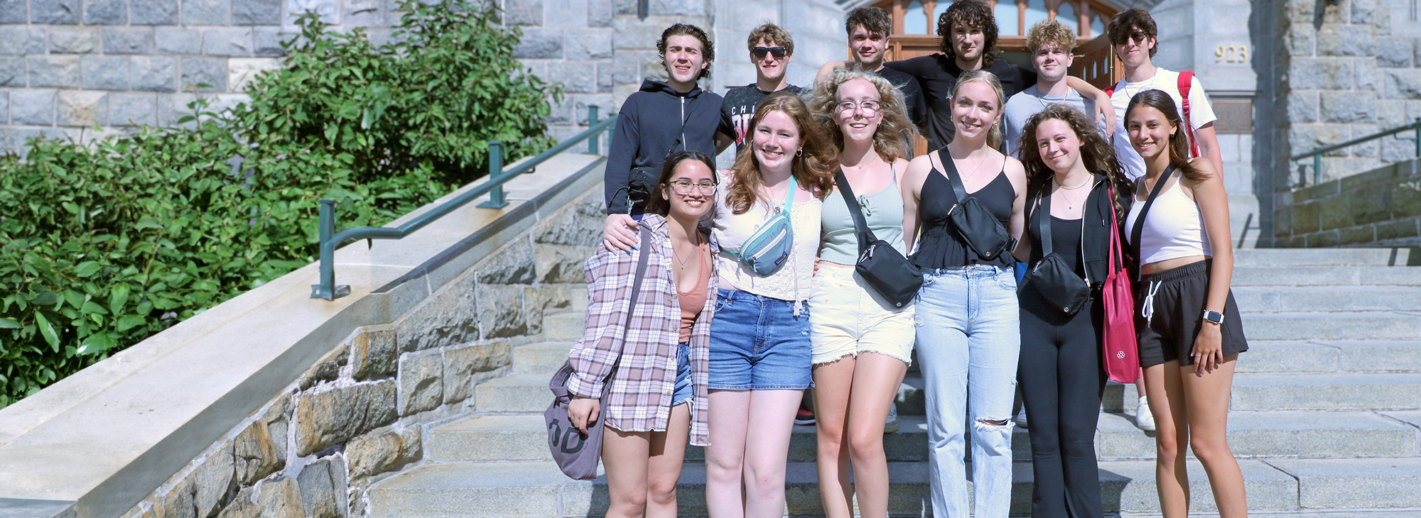 A group of students with arms over each other's shoulders,  standing on the stairs in front of McNally on a sunny day.