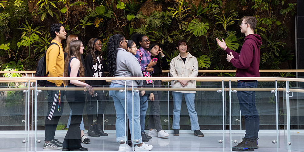 A current student gives a tour to future students. They are stopped in front of a wall of plants.