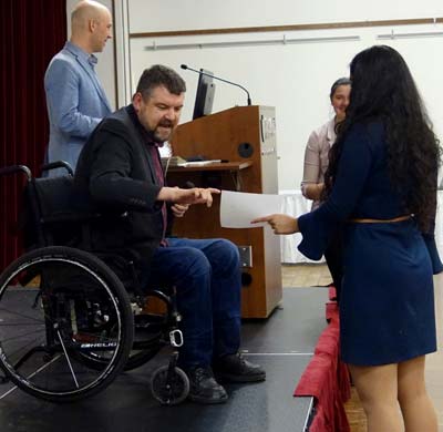 Man in wheelchair on a stage presenting an award to a female student who is stading on the floor next to the stage. A male speaker is in the background standing at a podium.